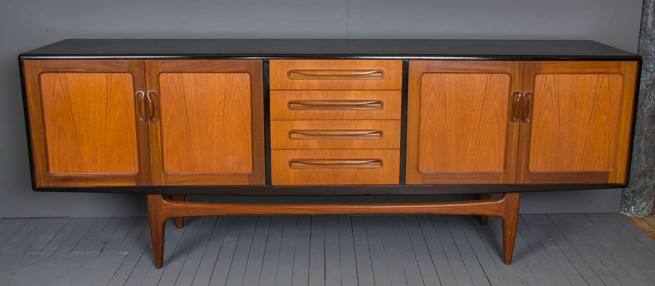 This excellent quality 1970s Teak sideboard features an ebonized finish top with solid wood panels, rounded corners and unusually shaped sculpted handles. The unit stands on 4 shaped legs joined by a stretcher bar in the front and the back. The