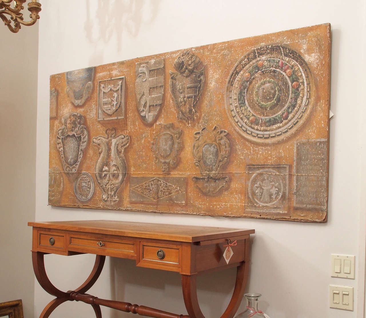 A large oil on canvas painting of architectural fragments, including shields and coats of arms, the fragments presented as if mounted on a painted plaster wall.  The canvas is of a coarse linen, with hand forged iron nails securing it to a highly