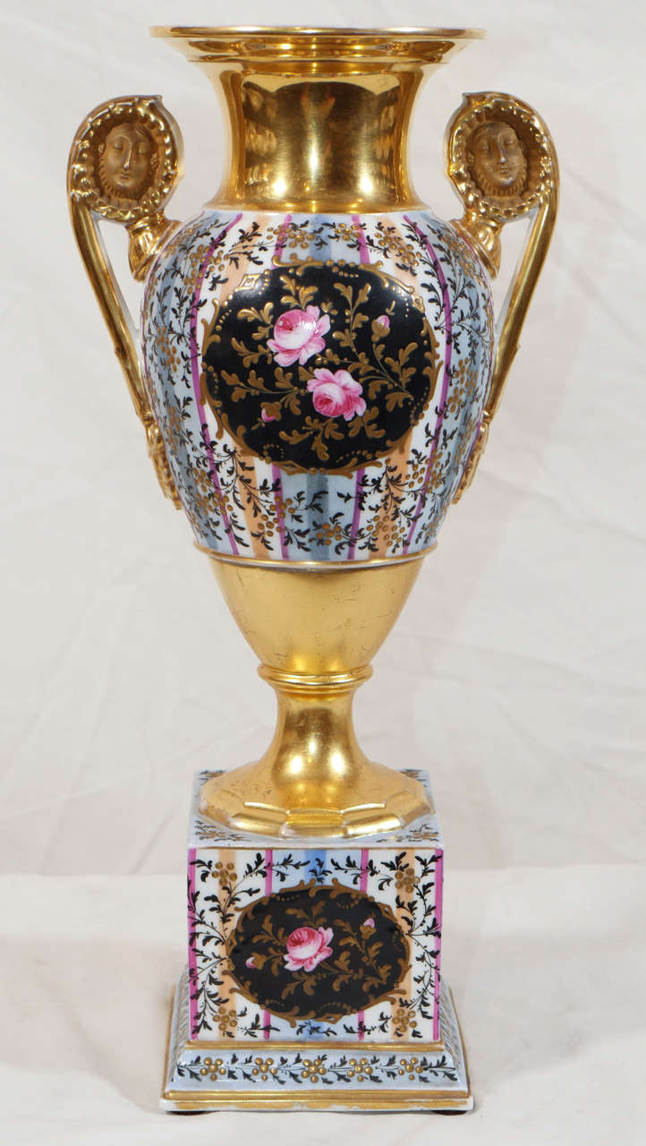 A pair of Louis XVI style, french, gilded vases painted in pale blue with stripes of lavender reserved with pink roses on a black ground.The vases stand on a square pedestal. They have mask handles. The neck and socle are brightly gilded.