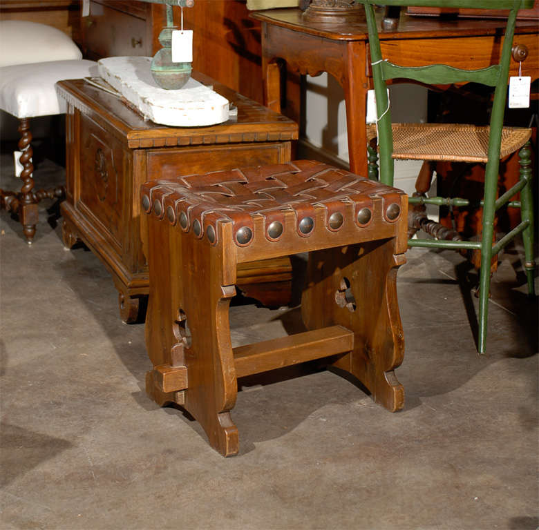 Taburete español de madera del siglo XX con tapa rectangular de piel tejida y cabezas de clavos de latón en el faldón. La base de caballete que lo sustenta presenta unos laterales en forma de lira con encantadores motivos de tréboles calados y un