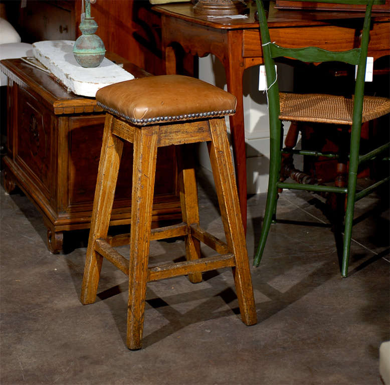 This 19th century Spanish wooden barstool features a comfortable rectangular seat with thick leather padding nicely outlined with brass nailheads. It rests on long splayed grain-painted legs with four footrests. The simple and sturdy design of this