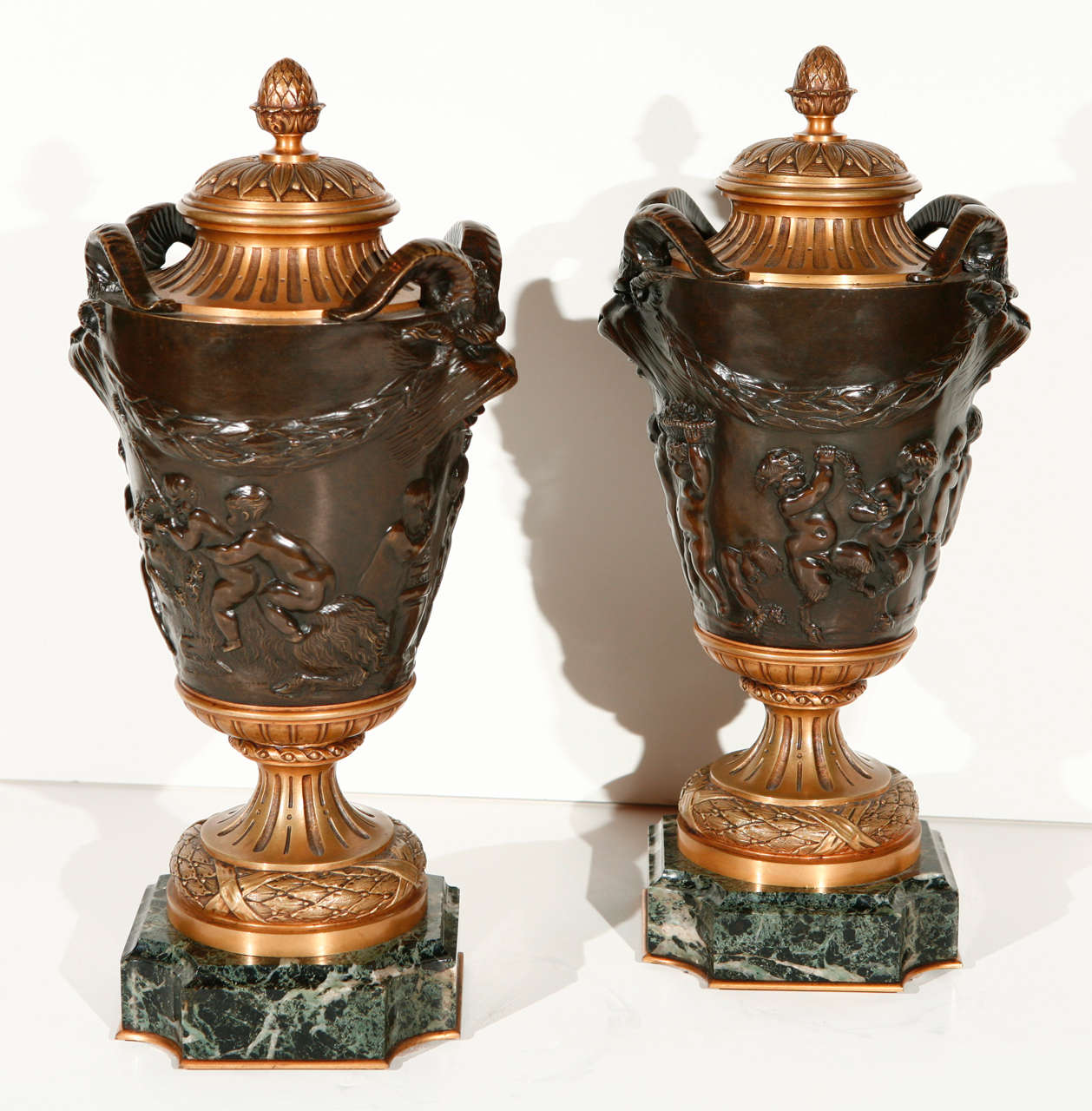 Pair of gilt and patinated bronze urns on marble bases featuring rams heads and putti.