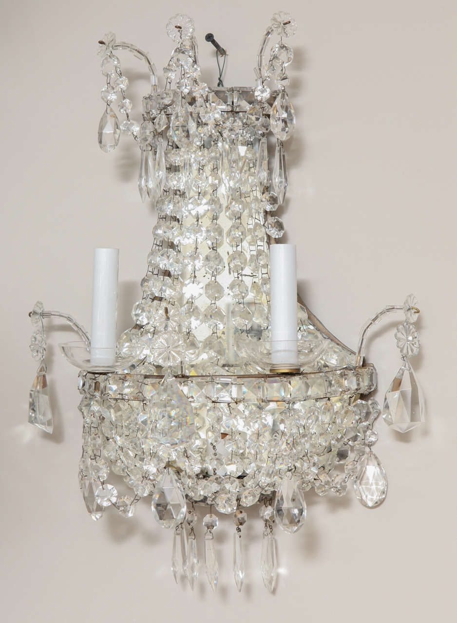 A pair of English Regency style two-light wall sconces with draped, faceted crystals of graduated size over frame having mirrored back. The interior of the sconce fitted with double candelabrum socket illuminating the mirror.