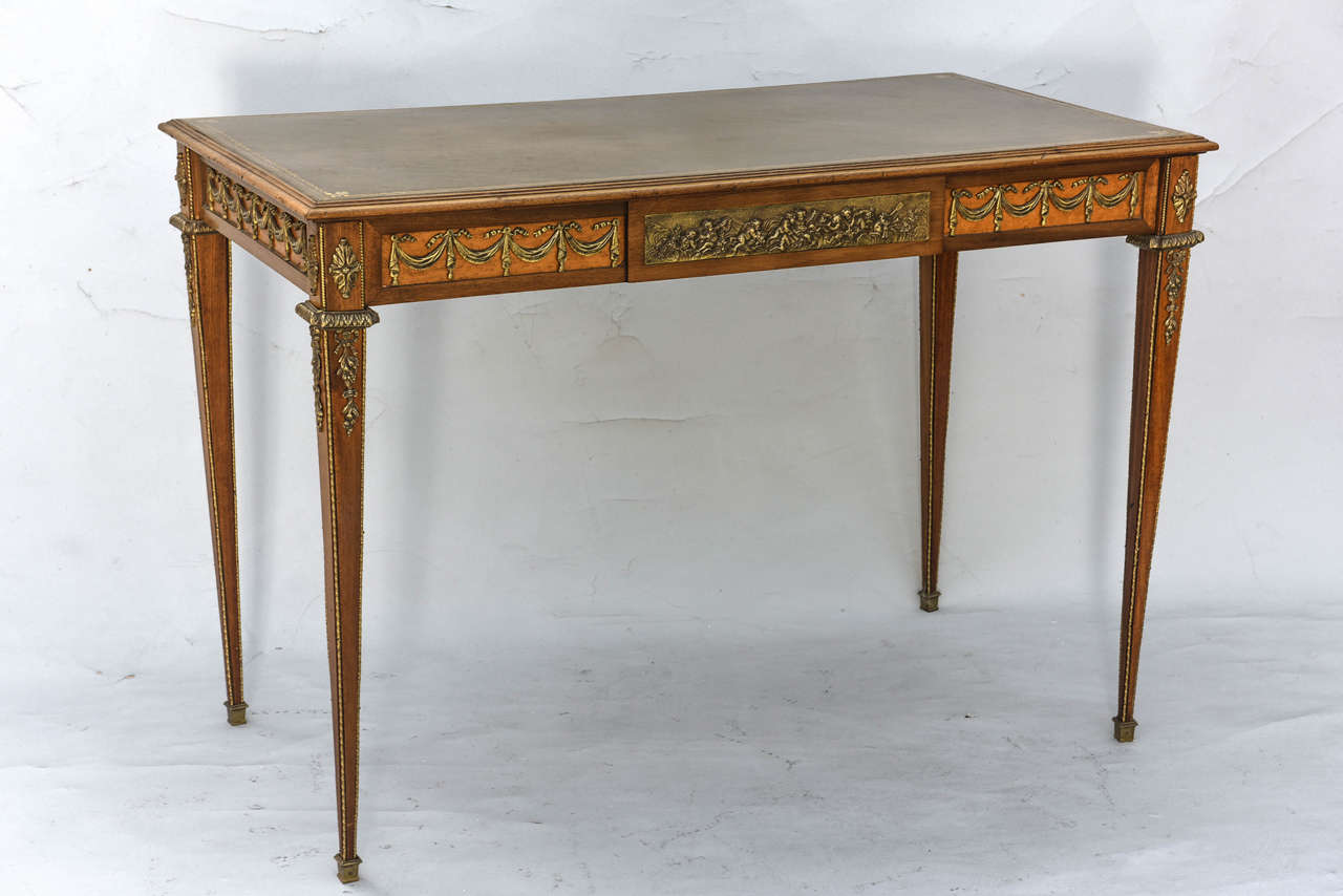 Writing table, of mahogany and satinwood, having embossed leather top, apron with elaborate bronze swags and drawer with inset bronze plaque of putti; raised on square tapering legs with additional bronze details and feet.