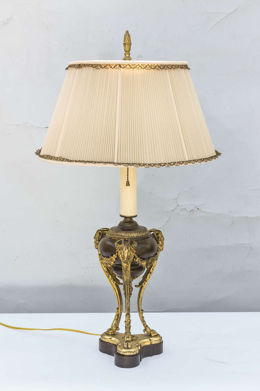 Athenienne table lamp, converted from an oil lamp,  the electrical fitting above a domed cover, the bulbous patinated bronze reservoir beneath a chased collar, descending to a  finial, on three gilt bronze tapering legs descending from ram's masks