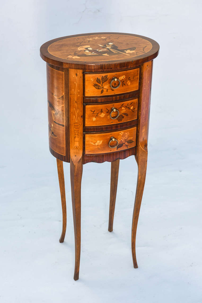 Marquetry decorated side table, of mahogany and mixed wood veneers, in the form of a petite commode, having an oval top on conforming base, with three stacked drawers, raised on delicate tapering cabriole legs, all its surfaces inlaid with
