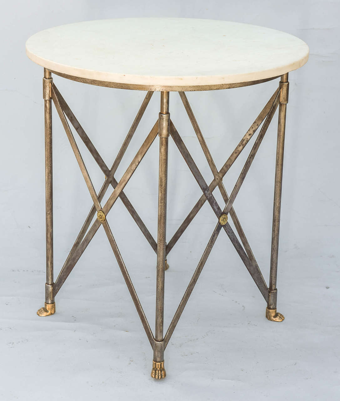 Occasional table, having a round top of white marble, on iron base, its four round legs joined by crossed vertical stretchers centered by a rosette, ending in brass paw feet.

Stock ID: D7777
