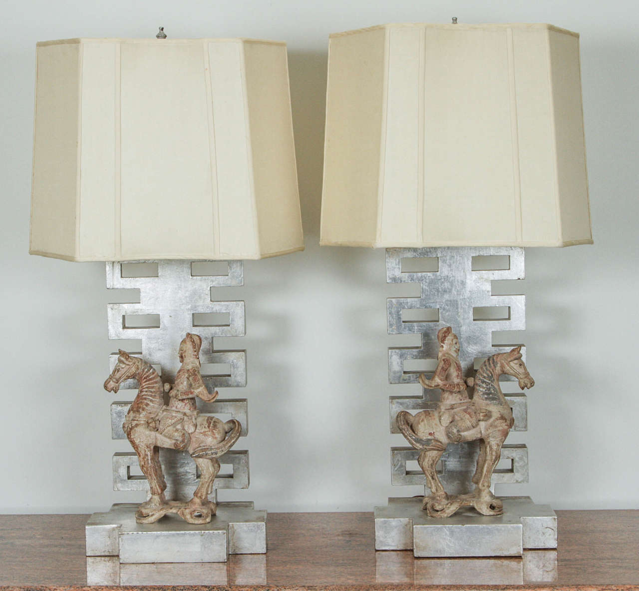 Gorgeous pair of table lamps by James Mont. They have silver leafed oriental style pedestals that support polychromed wooden figurines of equestrian Chinese warriors. The one sided fabric shades are original.