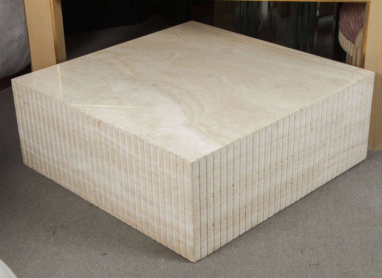 Cubic coffee table of polished travertine with carved vertical grooves on the sides. The heavy table is outfitted with wheels that are not visible and make it easily moveable.