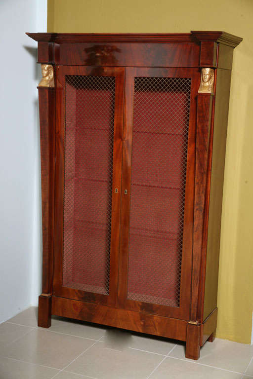 The whole in flame mahogany with Egyptian terms flanking the wire front cabinet doors- the interior later upholstered in red silk and fitted with later glass shelving.