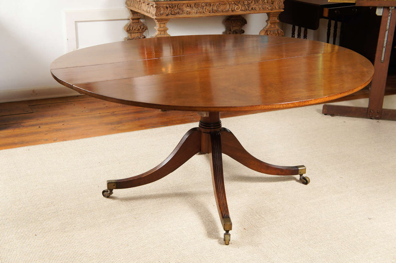 Nice English solid mahogany drop-leaf dining table. The top with one detachable leaf over a turned pedestal with four fluted downswept legs ending in brass cap toes and casters. This mechanical drop leaf allows the legs to be parallel or splayed to