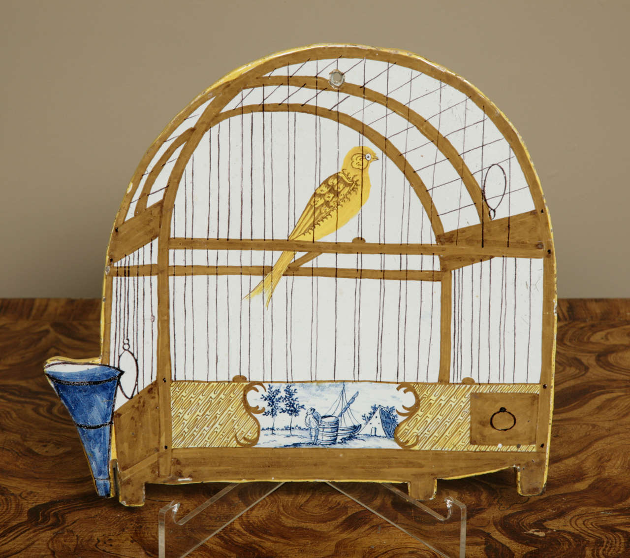 A charming and beautifully decorated and Delft birdcage plaque. Late 18th/early 19th century. (Dutch: Vogelkooiplaque). The arched plaque decorated is in, yellow, brown, black and blue with a perched bird, a water and seed feeder and a little Dutch