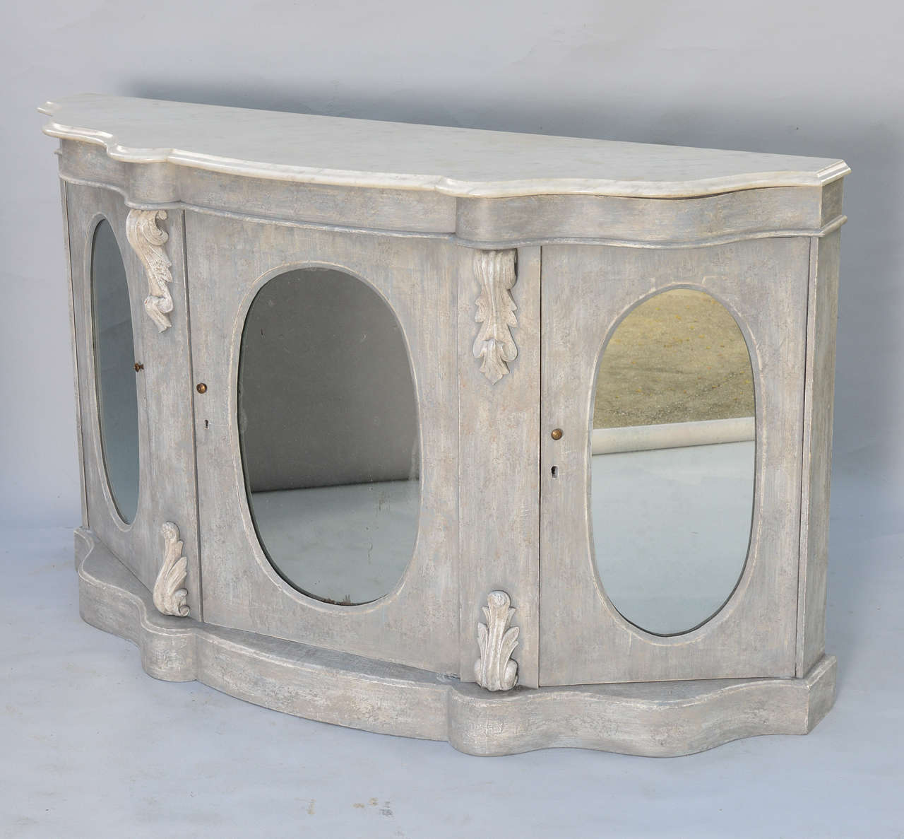 Buffet credenza or sideboard, with distressed painted finish, having a serpentine shape molded marble top, on conforming base with acanthus carved bracket details; its three cabinet doors each inset with oval mirror.