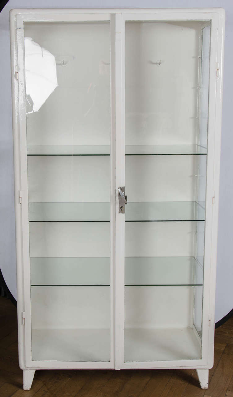 A large metal cabinet with shelves and hooks. This midcentury glazed industrial cabinet has a simple, utility style design with three glass shelves (which can be arranged at different heights) and a row of hooks running across the top. The cabinet