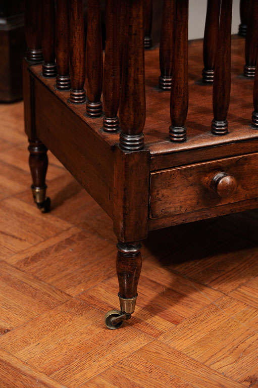 George III mahogany Canterbury with two rectangular divisions, turned splats above frieze drawer with ring turned tapering legs on brass feet and castors.