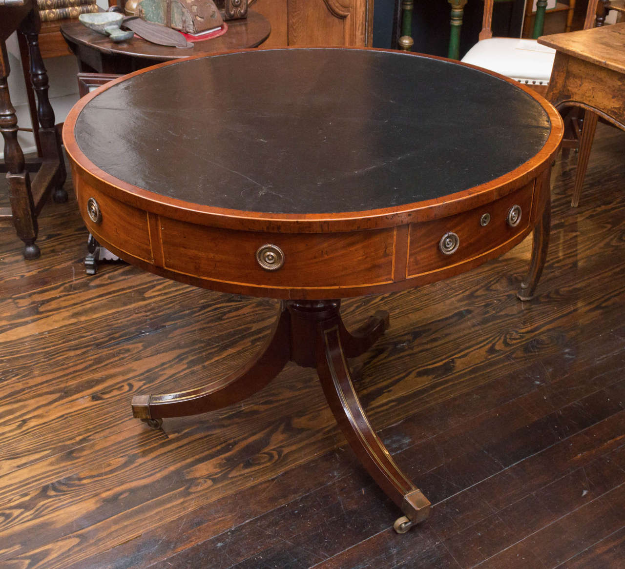 English Regency period drum table of mahogany with four drawers.  Old black leather top and inlaid brass pedestal. Brahma locks (no keys) and original casters. Excellent small size and solid construction. Sitting on the original brass casters.