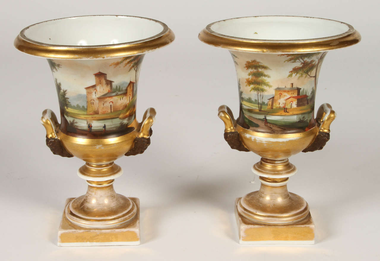 A very fine and charming pair of mid 19th century small porcelain de Paris urns. The urns are decorated with hand-painted landscapes and have gilt figural handles. The urns sit upon square gilt plinths. A little piece of Paris to liven up any space!