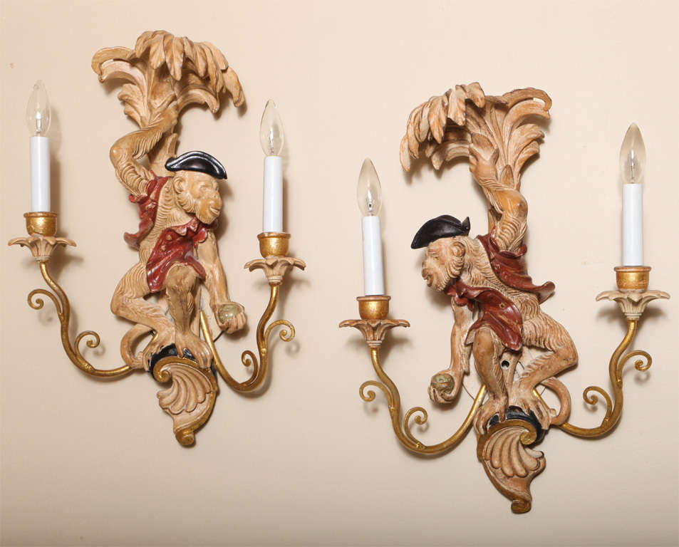 Opposing pair of whimsical wall sconces, of carved wood with painted and parcel gilt finish; each a dressed in vest and tricorn hat, standing on a scroll with a palm tree, two scrolling gilded iron candlearms terminate in leafy wooden bobeches. 