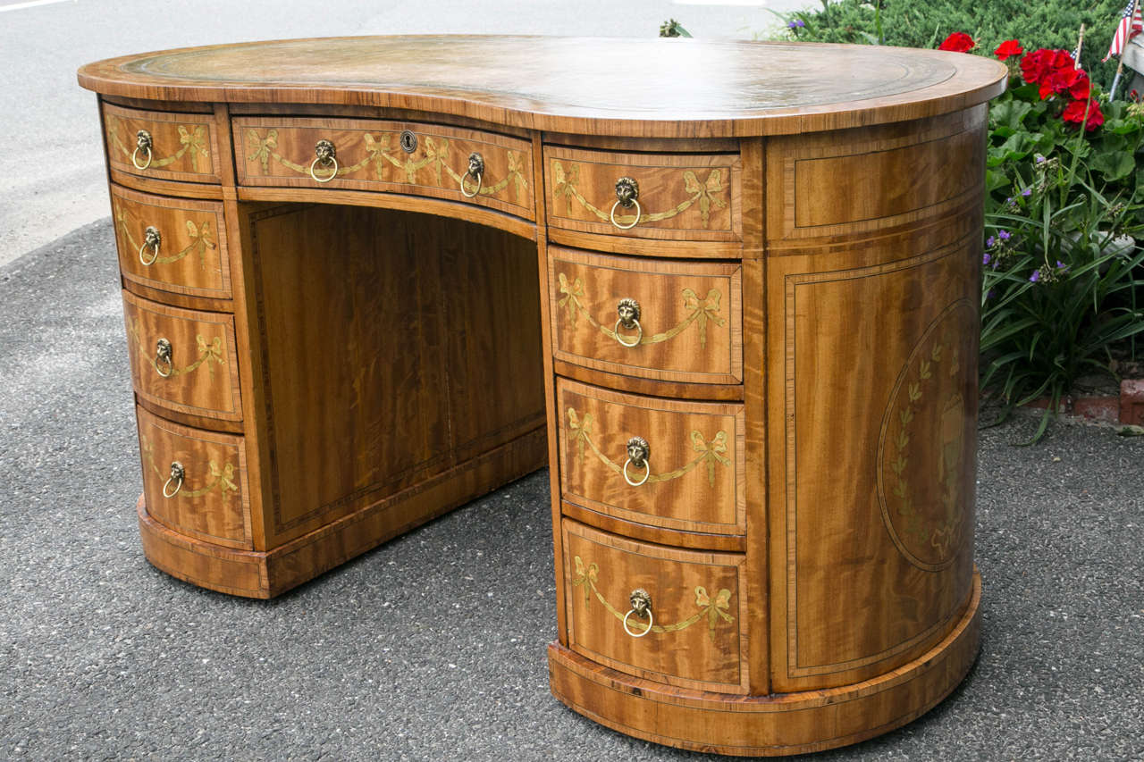 This desk, in satinwood, with inlays of sycamore, satinwood and mahogany has a leather top in pale olive.  The top has  a  tooled edge of two bands in black  surrounding a gold band.
A center drawer above the knee hole which measures 20 wide and 24