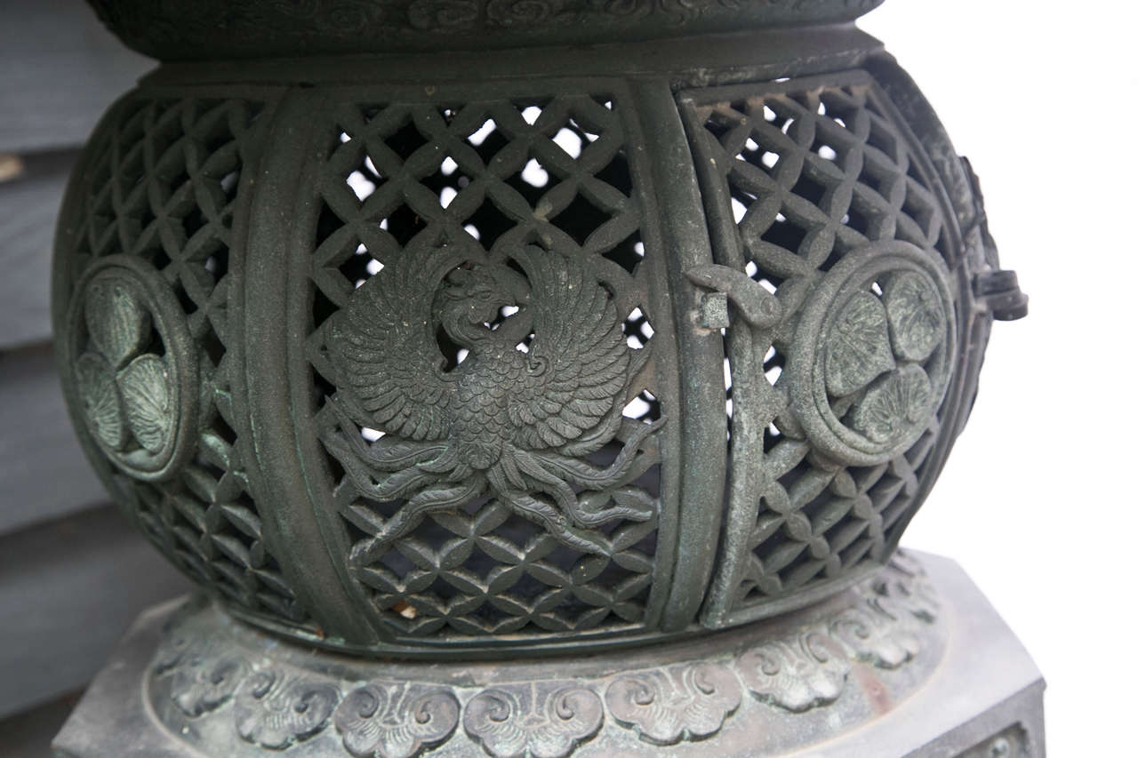 This bronze garden lantern in the archaic Japanese style is topped with a flaming orb, above a  typical form with  8 winged dragons emanating. The central lantern form is latticed and the  door, decorated with phoenix and florals, opens to place
