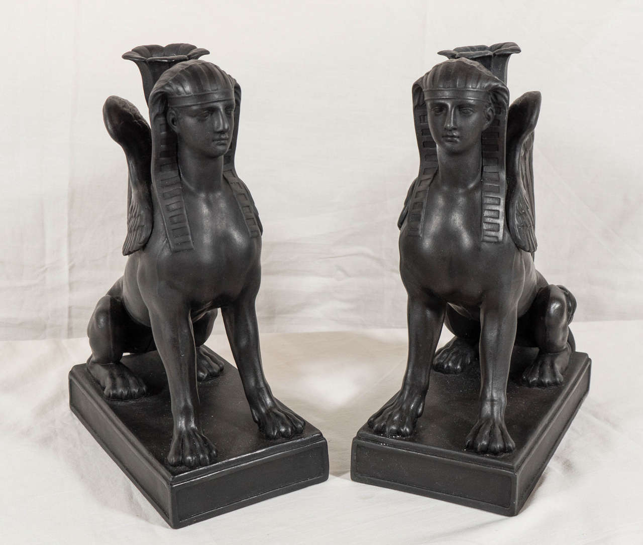 A pair of Wedgwood black basalt candlesticks in the form of sphinxes, each modeled seated upright atop a raised rectangular base with 'lotus' nozzle to hold candles. They have the head of a woman, the body of a lion and a pair of wings. This form