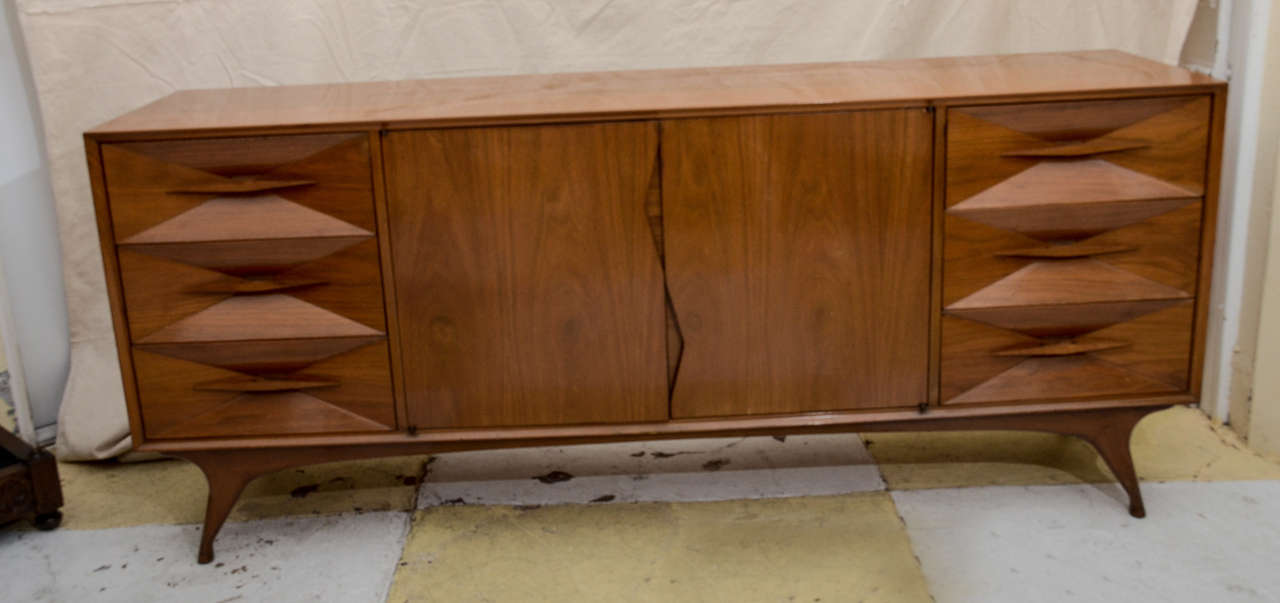1960 AMERICAN MID CENTURY MODERN CREDENZA-- SIDEBOARD RAISED ON 4 SPLAYED LEGS-- CENTERED BY 2 CUPBOARD DOORS WITH ZIG ZAG PROFILE THAT FUNCTIONS AS PULLS , BEHIND ARE 3 LINEN SLIDES-- 3 DRAWERS ON EACH SIDE WHOSE FRONTS FORM AN INVERTED PYRAMID