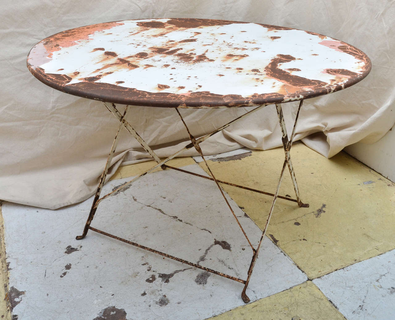 French old paint finish folding round iron garden table. Round distressed paint (white top coat with orange red under coat -- rusted areas and one area of bondo) top on X folding base with X support. The legs are joined by low stretchers, ending in