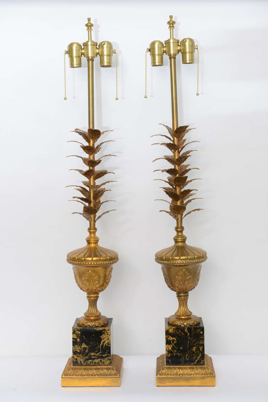 Featuring a double socket cluster, this pair of very elegant table lamps have a stem of gilt tole leaves rising from a column footed neoclassical urn with acanthus leaves over a black and gold faux marble painted metal base with a gilt acanthus leaf