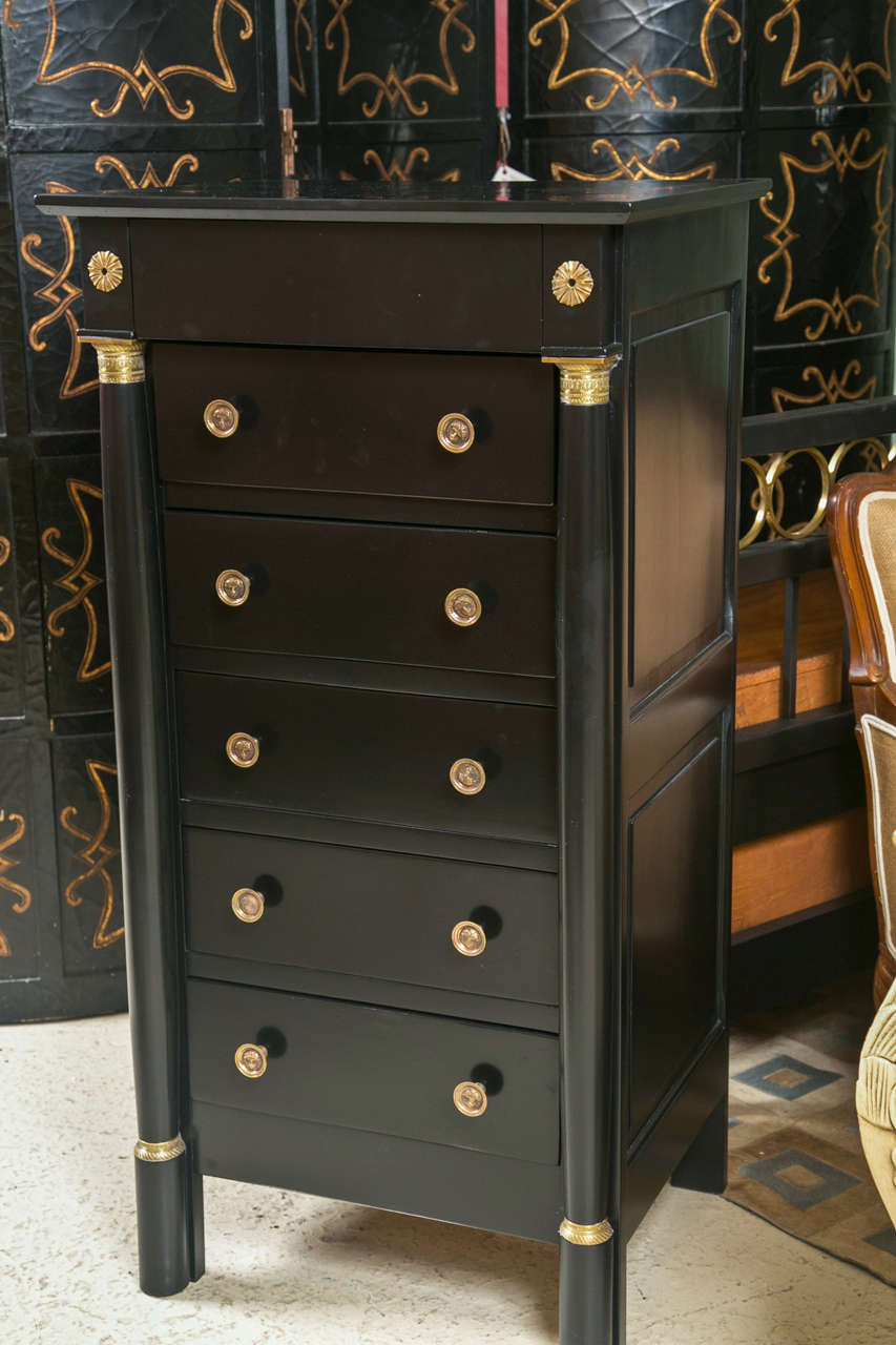 A French Empire style chest of drawers, possibly from the 1950s, overall ebonized, the top over a frieze decorated with brass pateras atop a conforming case fitted with five drawers, each having brass knob pulls, flanked by columnar upright on each