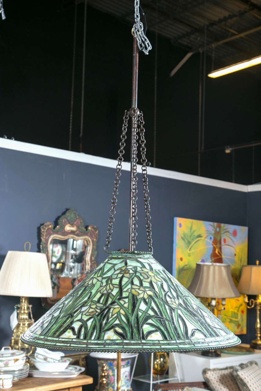 Lamp has Daffodil flowers in shades of green and yellow glass distributed throughout the cone shaped fixture. Long stems of yellow and green. At the bottom of the shade are three rows of glass and a beaded ring on the very bottom. Signed Tiffany