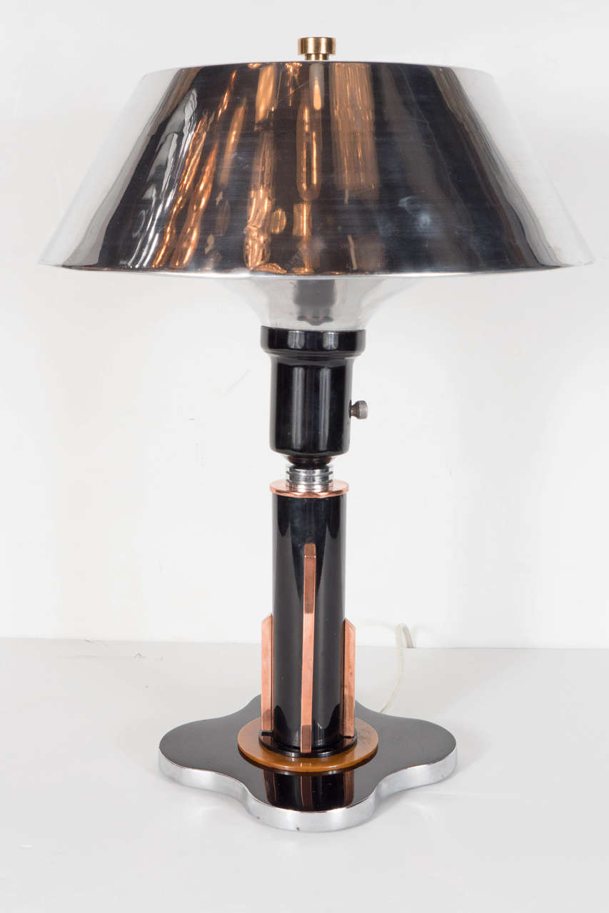 This beautiful Art Deco Machine age desk lamp features a black enamel base in quatrefoil design with a border of polished aluminum and a circular inset detail of amber bakelite and black enamel stem with accents of copper and chromed bandings and