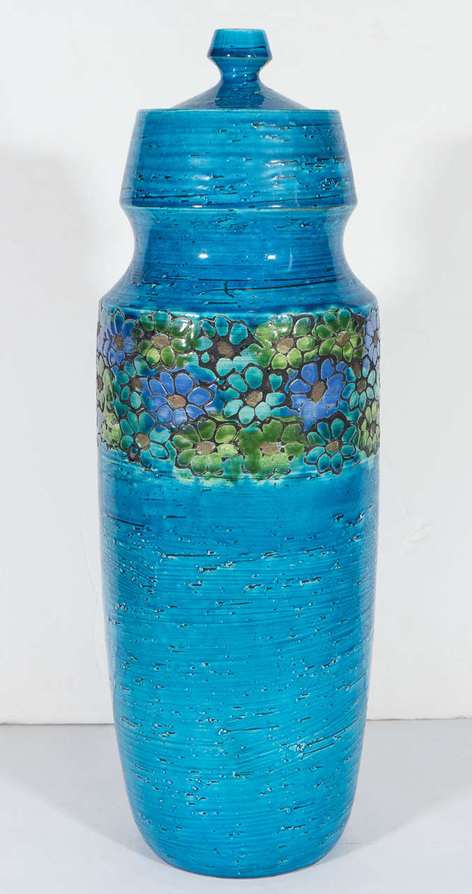 A tall, blue lidded jar in textured and glazed ceramic, produced in Italy, circa 1960s by Rosenthal Netter, decorated with incised flowers, glazed in blue, green and purple. Marked [67/7B] to the bottom of the jar, with the original label [Created
