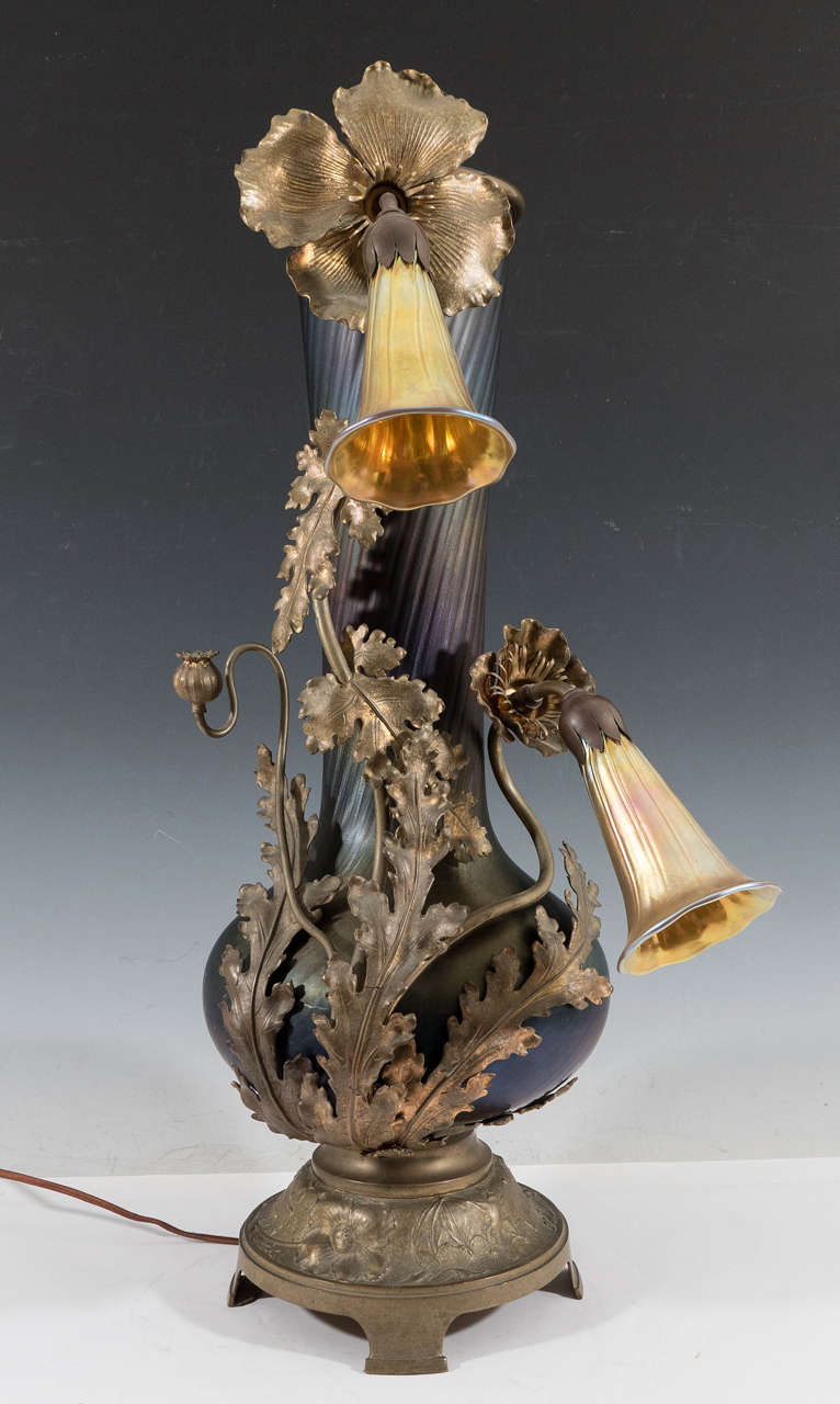  Austrian Art Nouveau table lamp, produced circa 1900s, in the form of a Deep Amethyst Glass Vase on a stylized bronze base, with a Bat and Poppy Flower and Buds Surround in bronze, supporting two iridescent Tiffany Glass Lily shaped glass light