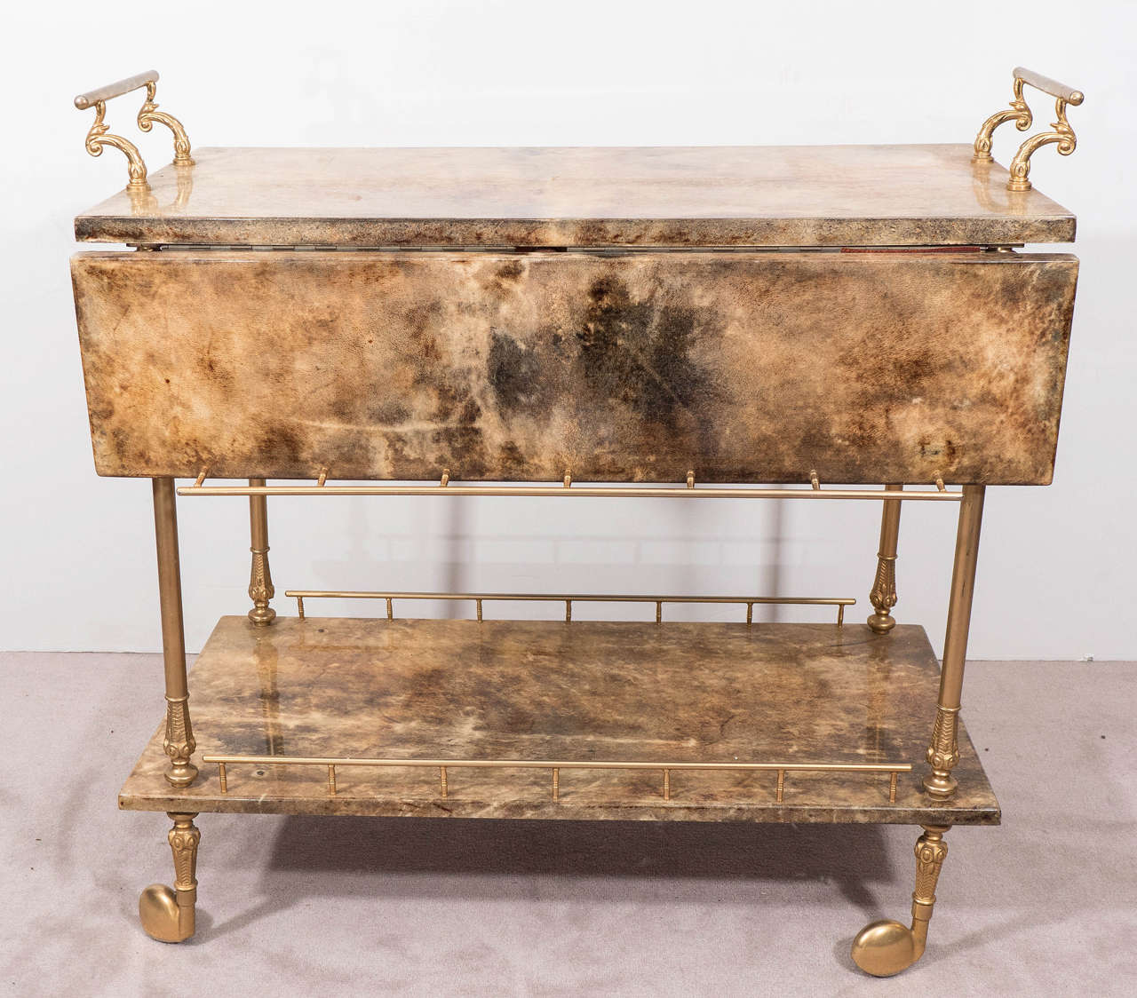 A vintage Italian two-tier bar cart, produced circa 1950's by designer Aldo Tura, veneered in lacquered goat skin, with scroll work handles, classically formed legs and casters in polished brass. Good vintage condition, consistent with age and use,