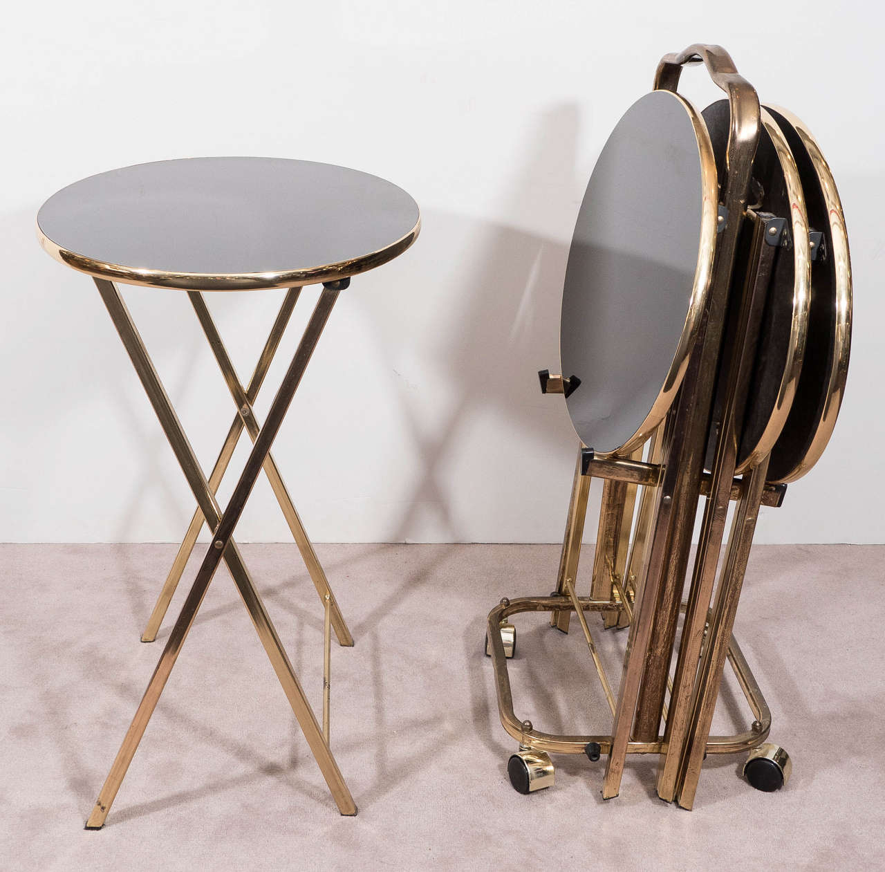 A vintage set of four dinner trays on a brass stand with casters, produced Midcentury in the Hollywood Regency style, each with glass tops in black, inset in brass frames on folding X-form legs and spreaders. Good vintage condition with age