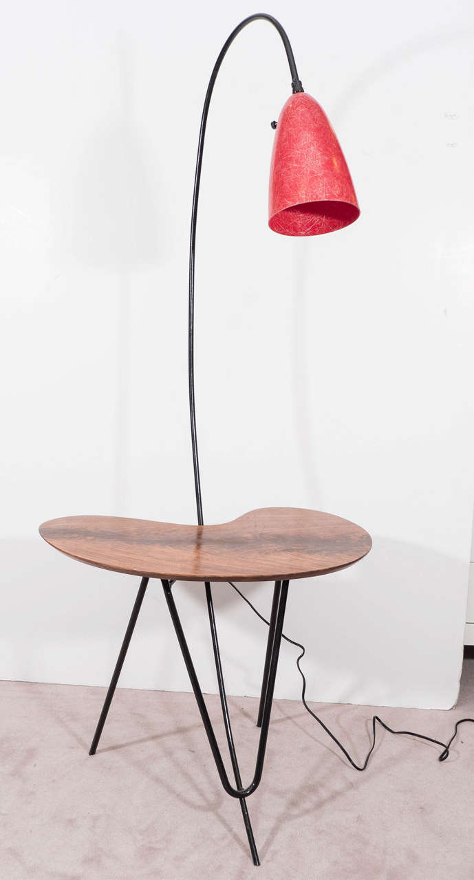 A vintage floor lamp, produced, circa 1950s in the style of Swedish designer Greta Grossman, with red Fiberglass lamp shade, affixed to a curved stem on hairpin legs in brass with black enamel, inset by a wooden palette shapes table. Wiring and