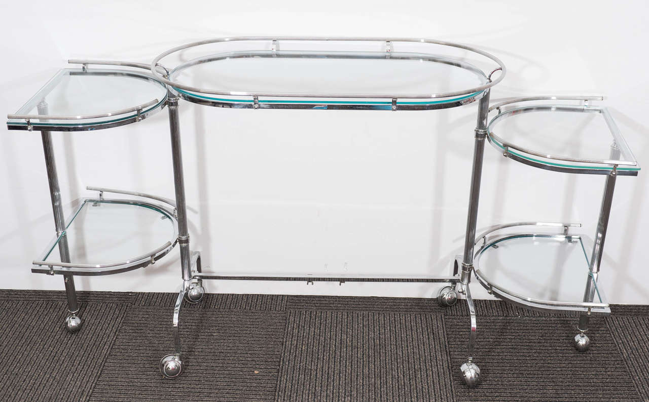A vintage, five-tier bar cart, with chrome frame, inset with glass shelves, able to swivel and expand outwards, with reeded gallery rails and legs, on six casters. Very good vintage condition, consistent with age and use.

Width fully extended: