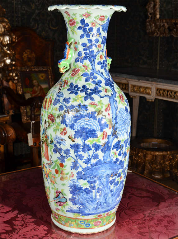 An oversized vase, celadon background with trees, butterflies, flowers and people in relief on blues, reds, greens and yellows with gilt accents and Foo dogs at neck.