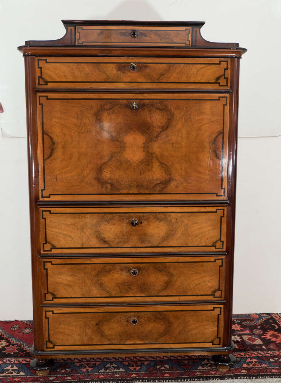 This tall, slim secretaire is crafted of solid walnut and fir, and exquisitely veneered and inlaid with French Walnut and ebonized birch. The secretaire features five locking drawers surrounding a central drop-faced writing surface. When open, the