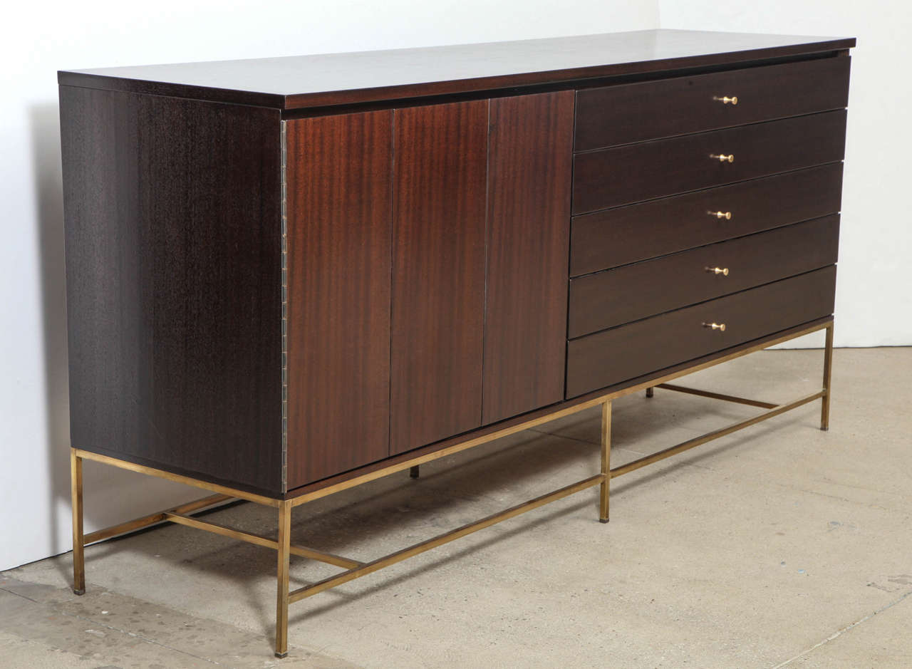 Mid Century classic chocolate brown stained Mahogany credenza with 5 drawers and a side compartment with trifold doors which conceals an adjustable shelf. Credenza rests on a brass stretcher base.