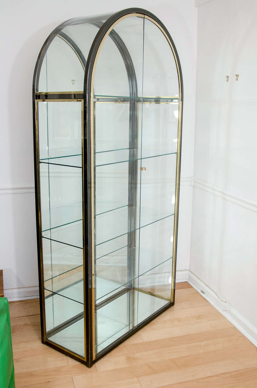 Elegant arched vitrine in brass and gunmetal glazed steel. Features four glass shelves plus mirrored back and base. Signed with DIA sticker.