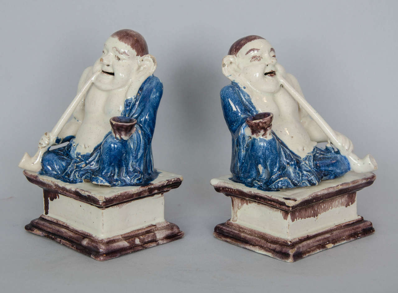 A pair of northern European faience pagoda figures (Pu-Tai), probably German, first quarter of the 18th century.
Each corpulent male figure modelled seated smoking a pipe, wearing a blue robe and holding a manganese tea bowl, on a stepped