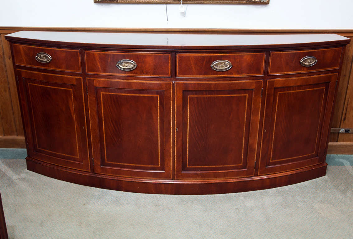 Made for us in England, this mahogany bow front server/credenza features four drawers over four recessed panel doors. The top and front have a narrow string of satinwood and a wider satinwood band. Oval brasses are used on the drawers; doors are