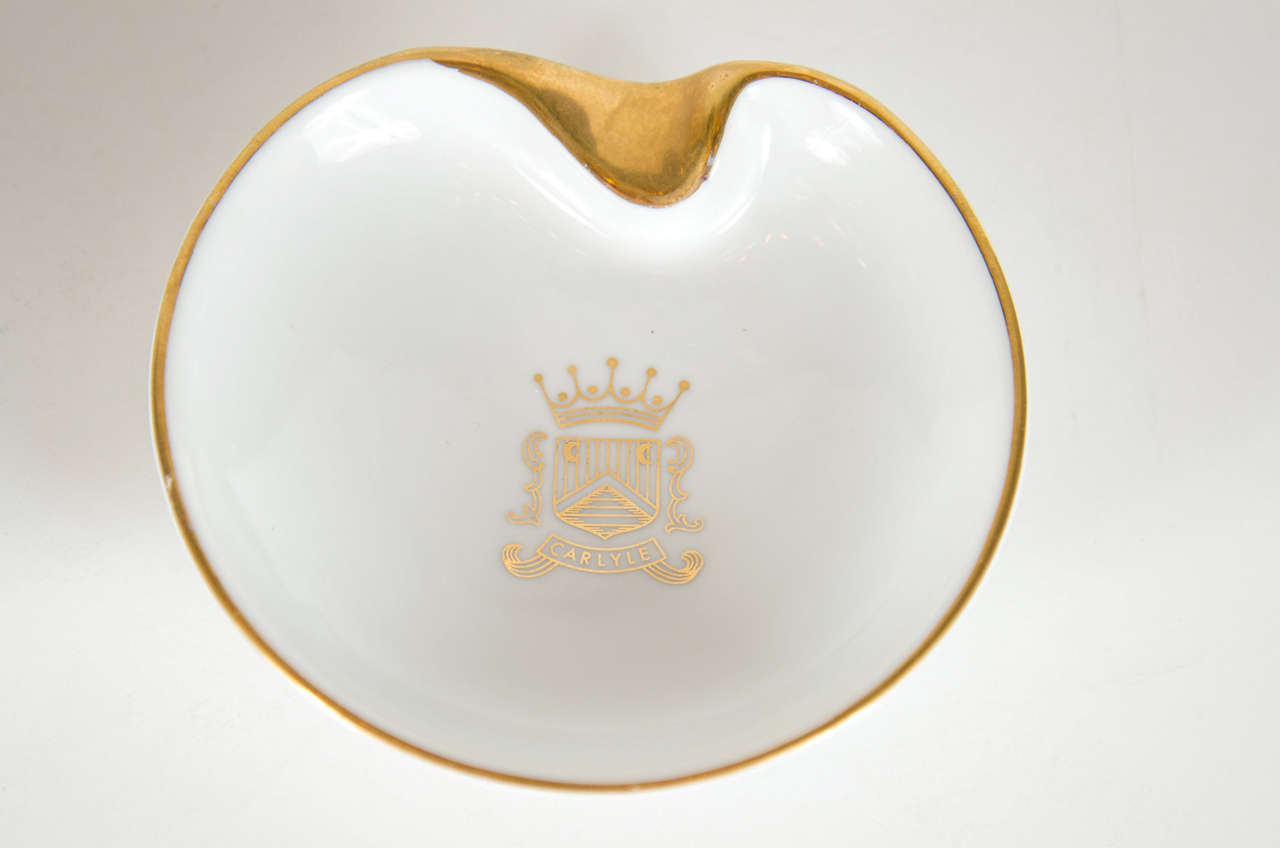 This elegant ashtray features white porcelain with a 24k gilt detailing design. It bears the name Carlyle and their crest in the center and it's a modernist organic form.