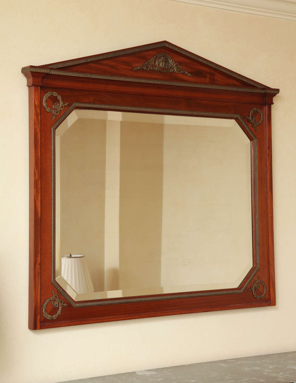 The beveled mirror plate with canted corners within a rectangular frame applied with laurel wreath mounts and foliate banding surmounted by a triangular pediment centered by a Classical mask.