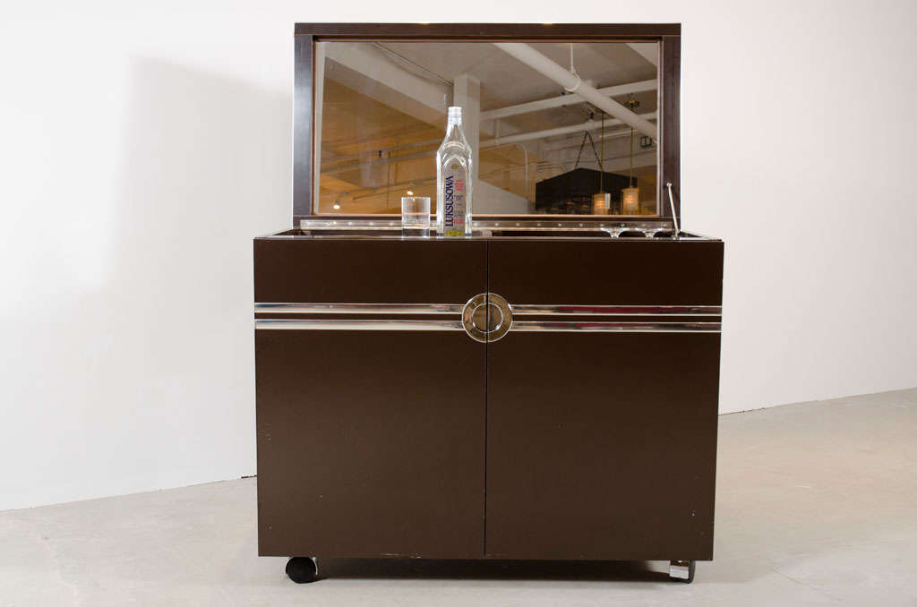 A chic chocolate colored laminate and polished chrome dry bar in a two-door form on casters with lift-up top that is mirrored on the inside and conceals a removable white plastic ice bucket, doors conceal one drawer and open storage, original brown