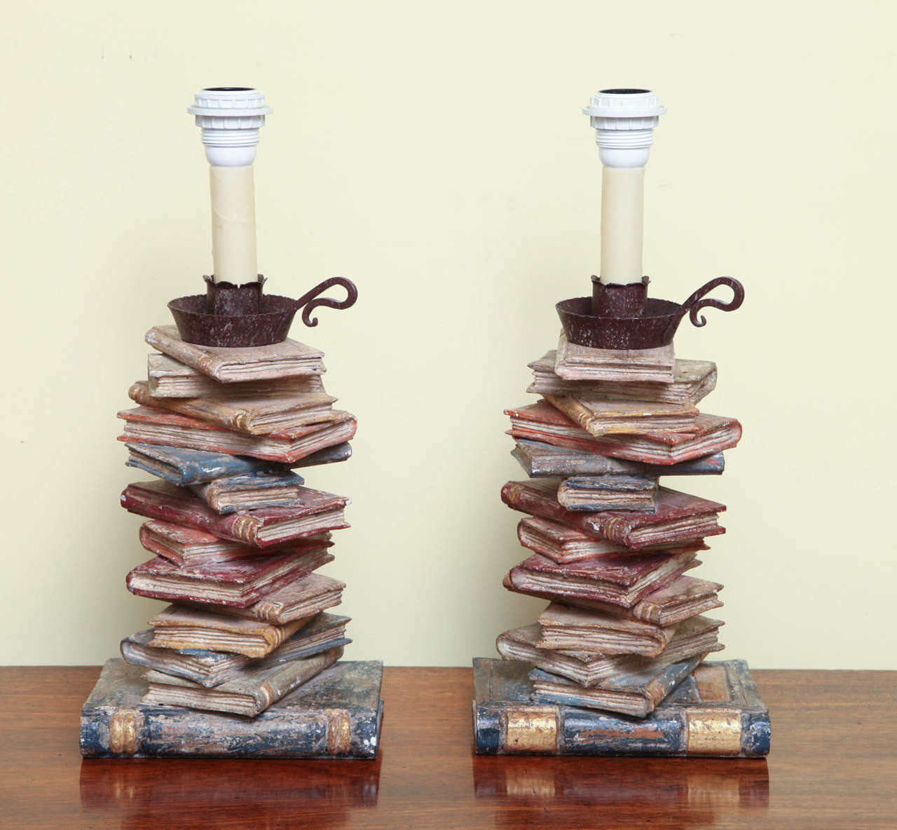 An amusing pair of polychrome and parcel-gilt carved wooden candlesticks in the form of stacks of books with a forged iron candlestick with holder to the top, now electrified as lamps, Italian, circa 1920.