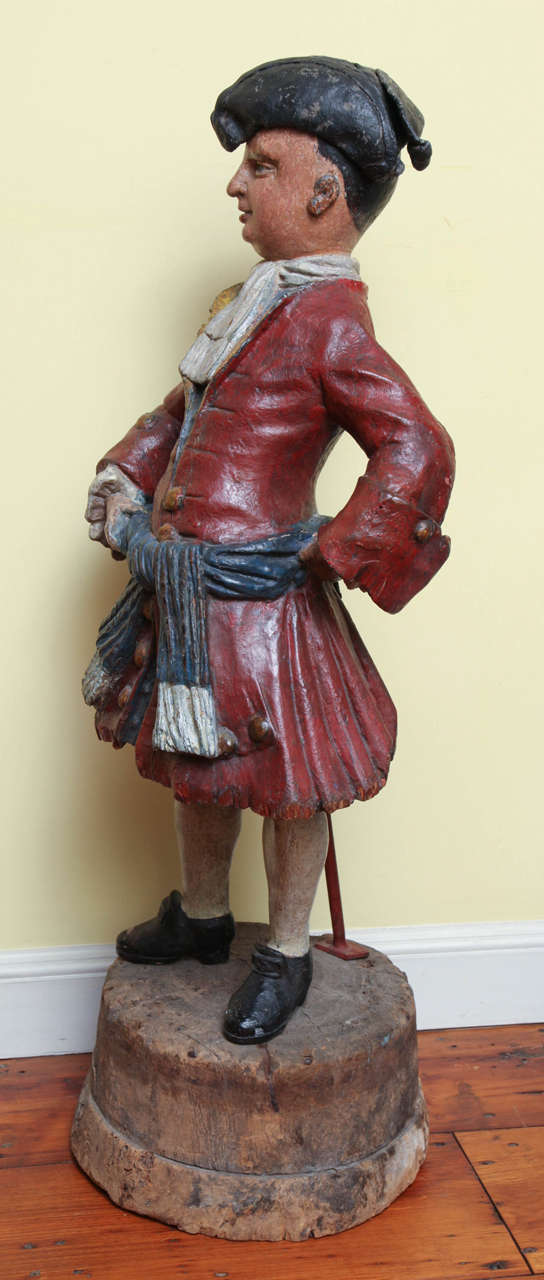 Extremely rare and exceptional George III carved wood polychrome store figure of a man in mid-18th century costume, sporting a solid lead tri-corn hat and lead ears and wearing a red coat with blue waistcoat, a prominent white sash tied at the neck