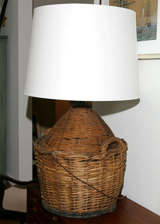 Green glass wine jug altered into a lamp with white shade, covered with hand woven wicker on a solid slatted wooden base