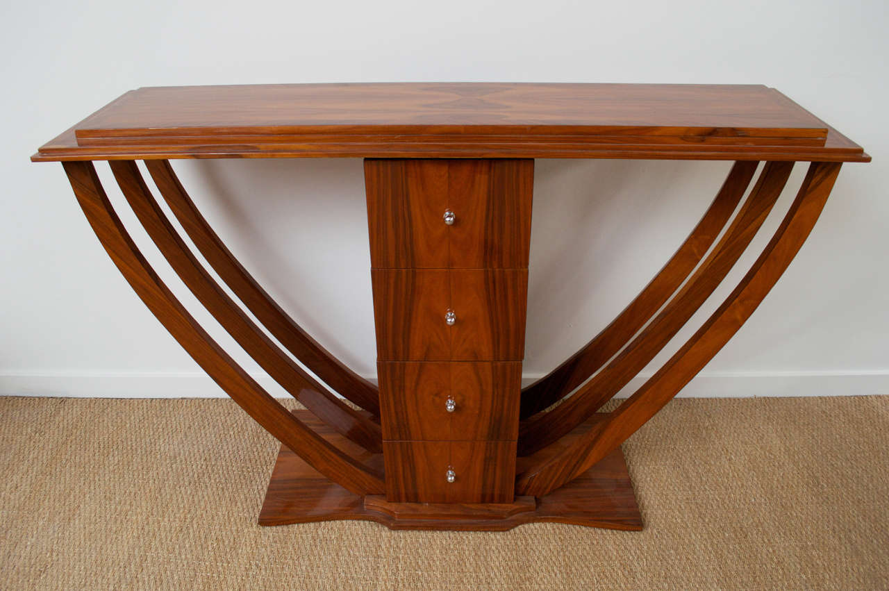 An art deco console table of varnished rosewood, and metal drawer pulls. Features a post of four drawers, and flared, rib-like legs.